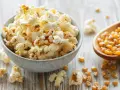 Schüssel mit frisch zubereitetem Popcorn im Topf, daneben eine Holzschaufel voller Maiskörner.