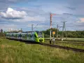 Zielony pociąg hybrydowy PKP jedzie po torach wśród zielonych pól. W tle las i chmury.