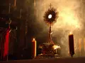 Złota monstrancja z hostią, otoczona płonącymi świecami i dymem, tworzy atmosferę głębokiej adoracji.