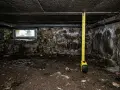 Mètre-ruban dans un vide sanitaire sombre, mesurant la hauteur sous le plafond en béton. Murs de pierre humides et sol terreux.