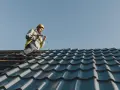 Murarz w kasku i kamizelce pracujący wkrętarką na dachu z niebieskiej dachówki.