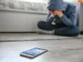Osoba w kapturze siedzi na podłodze, ukrywając twarz w dłoniach. Obok leży telefon, symbolizujący izolację i dystymię.