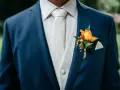 Elegancki, granatowy garnitur na wesele z białym, wzorzystym kamizelką i krawatem. Na klapie marynarki ozdobna butonierka z żółtą różą.