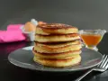 Pila di pancake senza latte, dorati e soffici, con sciroppo d'acero e uova sullo sfondo.