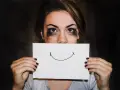 Dziewczyna z rozmazanym makijażem ukrywa usta za kartką z uśmiechem. Trudna samoregulacja emocji.