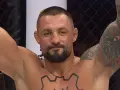 Mateusz Janur, wojownik MMA, z podniesionymi rękami i tatuażami na ramionach i klatce piersiowej, świętuje zwycięstwo.
