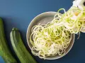 Espaguetis con calabacín recién hechos, listos para servir. Dos calabacines verdes reposan al lado de un plato lleno de fideos de calabacín.