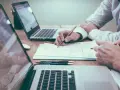 Dos personas revisan documentos y trabajan en laptops. Un modelo 200 cuando se presenta, con gráficos y notas.