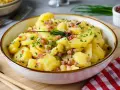Schüssel mit warmem Kartoffelsalat, garniert mit Speckwürfeln und Schnittlauch. Ein Klassiker, der immer schmeckt!