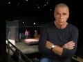 Dawid Głowacki, reżyser teatralny, z rękami skrzyżowanymi na piersi, na tle sceny z łóżkiem i ludźmi.