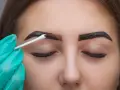 Ręka w rękawiczce próbuje ratować nieudaną hennę brwi, usuwając nadmiar ciemnej pasty wacikiem.