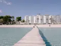Drewniany pomost prowadzi do piaszczystej plaży Playa de Muro z białym hotelem i palmami w tle.