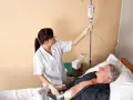 Pielęgniarka podłącza kroplówkę do pacjenta leżącego w łóżku. Widać rurkę od PEG.