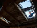 Mężczyzna montuje nowe okno dachowe. Widać drewniane więźby dachowe i fragmenty starego okna.