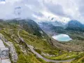 Strada tortuosa sul Passo San Lugano, con lago alpino azzurro e montagne nebbiose sullo sfondo.