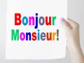 Una mano sostiene un cartel con "Bonjour Monsieur!" escrito en letras de colores.