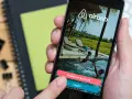 Dłoń trzyma smartfon z aplikacją Airbnb. Czy Airbnb jest bezpieczne? Na ekranie widać basen i dzieci, przyciski "Create an Account" i "Log In".