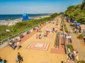 Tętniąca życiem ustka promenada w słoneczny dzień. Ludzie spacerują, odpoczywają na ławkach, a w tle widać plażę pełną turystów i morze.