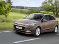 Brązowy Hyundai i20 na drodze wśród zieleni. Kierowca uśmiecha się, co może sugerować pozytywne hyundai i20 opinie.