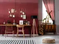 Eleganckie biurko z drewnianym blatem i złotymi krzesłami na tle ściany w kolorze marsala. Wygodny fotel i lampa stojąca dopełniają przytulną przestrzeń.