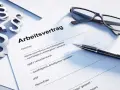 Drei Paragraphen-Symbole, eine Brille und ein Stift liegen auf einem Dokument mit der Überschrift "Arbeitsvertrag". Dies unterstreicht, dass ein Arbeitsvertrag schriftlich sein muss.