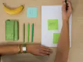 Une main écrit "STATUT" sur un post-it vert, à côté d'un carnet, de stylos et d'une banane. L'acronyme "EI" sur un post-it bleu suggère une **entreprise individuelle**.