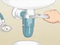 A hand uses a wrench to loosen a sink trap, a step in how to clean a sink drain. Water drains into a bucket below.