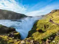 Potężny wodospad Gullfoss w Islandii, z mgiełką unoszącą się nad kaskadami. Ścieżka turystyczna wije się po zielonym zboczu.