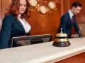 Recepcjonistka i recepcjonista za ladą hotelową. Dzwonek do obsługi gości, który pomoże dowiedzieć się, ile kosztuje doba hotelowa.