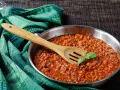 Un saporito ragù di cinghiale in padella, pronto per condire la pasta. Un cucchiaio di legno e una fogliolina di basilico completano la scena.