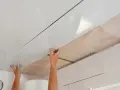 Pose de lambris PVC au plafond avec clips. Les mains ajustent une lame blanche brillante sur une structure, révélant un treillis blanc en dessous.