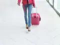 Kobieta w różowej kurtce i jeansach ciągnie różową walizkę, idealną jako wizzair bagaż podręczny, po białej, geometrycznej posadzce.