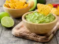 Pyszne guacamole w drewnianej misce, idealne do maczania nachos. Obok limonki i miska pełna chipsów. Z czym jeść guacamole? Z nachos!