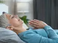 Frau liegt entspannt im Bett, Hände vor der Brust gefaltet. Sie praktiziert eine Technik, die wirkt Meditation im Schlaf.