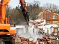 Pomarańczowa koparka niszczy dom, unosząc tumany kurzu i gruzu.
