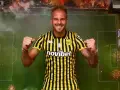 Uros Racic, con su nueva camiseta a rayas amarillas y negras, celebra con los puños en alto. El fondo simula un campo de fútbol con bengalas.