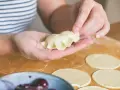 Ręce formują pieroga z ciasta na pierogi bez jajka. Na blacie leżą okrągłe placki z ciasta i miseczka z wiśniami.