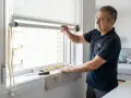 Técnico instalando motor para persiana, con herramientas y nivel a la vista. Un móvil muestra la interfaz de control.