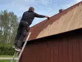 Man på en stege målar plank på ett tak med brun färg.