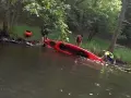Ludzie wyciągają czerwoną kajak z wody. W tle widać drzewa i drugą czerwoną łódkę. Woda jest ciemna, jakby to było jezioro smołowe.