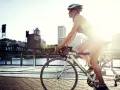 A female cyclist enjoys a sunny ride along a waterfront path with a city skyline in the background.