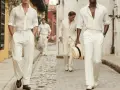 Uomini con un elegante outfit uomo pantaloni bianchi a righe, camicie leggere e cappello di paglia, passeggiano su un vicolo acciottolato.