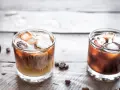 Dwa drinki na drewnianym stole: jeden to kremowy White Russian, drugi to ciemny Black Russian.