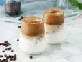 Två glas med krämigt dalgona kaffe med en fluffig kaffetopping över mjölk och is. Kaffebönor sprider sig på marmorbordet.