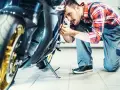 Mechanik w kratkę wykonuje przegląd motocykla, pochylając się nad jego przednią częścią.