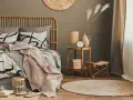 Dormitorio con cama de ratán, ropa de cama beige y gris, manta rosa, mesita auxiliar de madera y alfombra redonda.