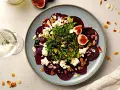Rote Bete Carpaccio mit Feigen, Ziegenkäse und Mandelsplittern auf einem blauen Teller, daneben ein Glas Weißwein.