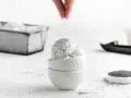 Eine Hand streut Mohn über cremiges Mohn Eis in weißen Schalen. Ein Löffel und eine Schale Mohnsamen liegen daneben.