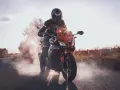 Motocyklista w kasku i kombinezonie na pomarańczowym motocyklu Barton Blade, z opon dymiących na drodze.