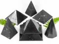 Seis pirámides de piedra shungita, algunas con vetas blancas, dispuestas sobre un fondo blanco con hojas verdes.